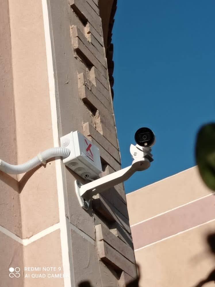 Small black security camera on a white mount attached to a textured beige building wall.