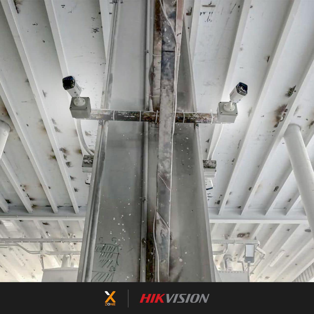 Two white security cameras mounted on a metal beam under a white industrial ceiling.