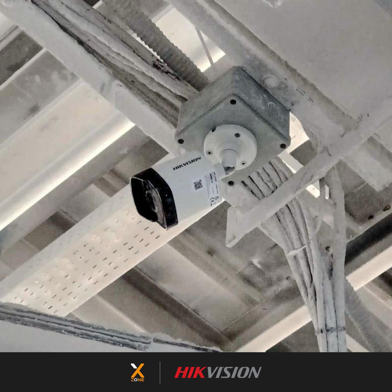 White Hikvision bullet camera mounted on a grey junction box against a white industrial ceiling.