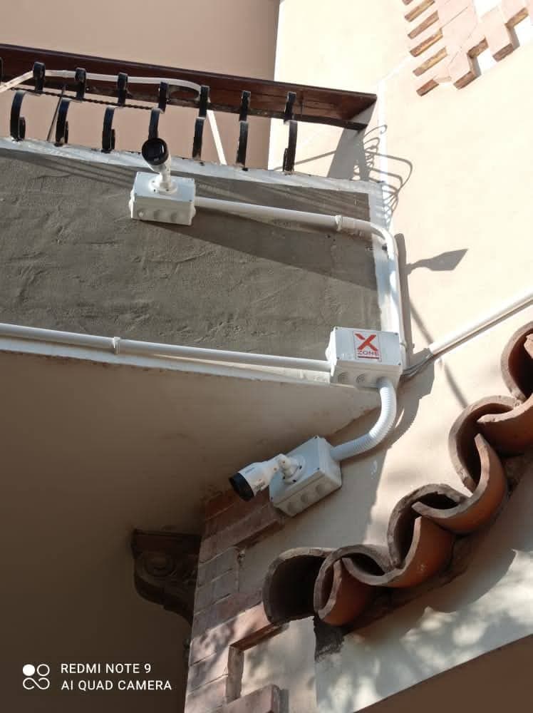 Two white security cameras mounted on a building's exterior wall and balcony with white conduit.