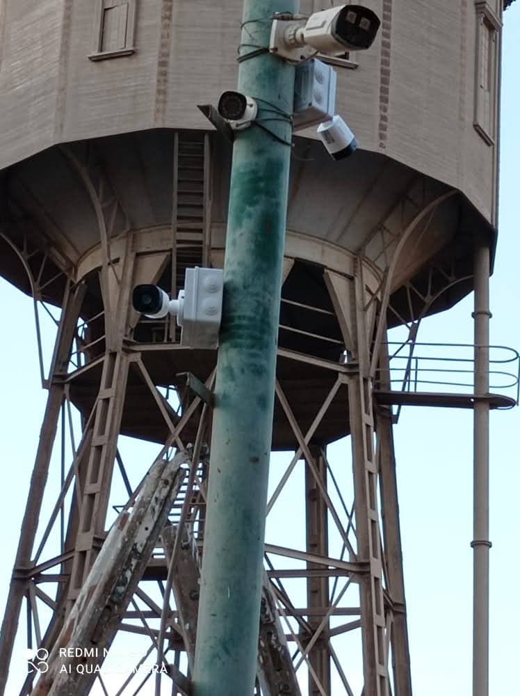 Multiple security cameras mounted on a tall pole with a large wooden water tower behind.