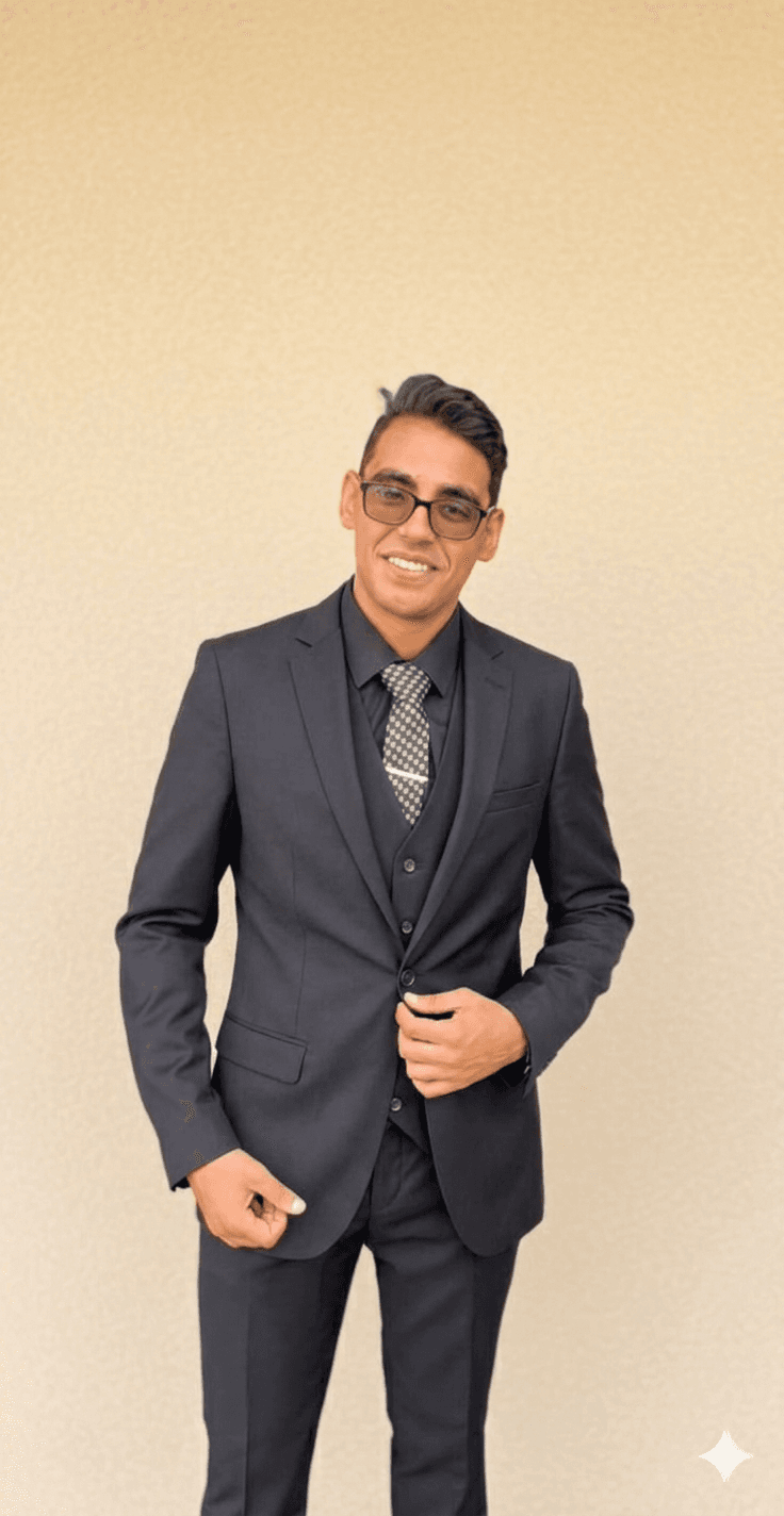 Smiling man in a black three-piece suit, glasses, and patterned tie against beige wall.