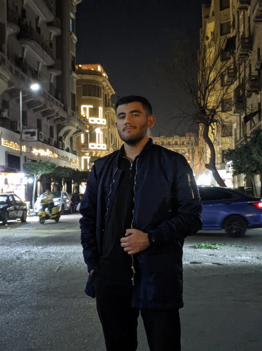 A bearded man in a navy bomber jacket stands on a city street at night.