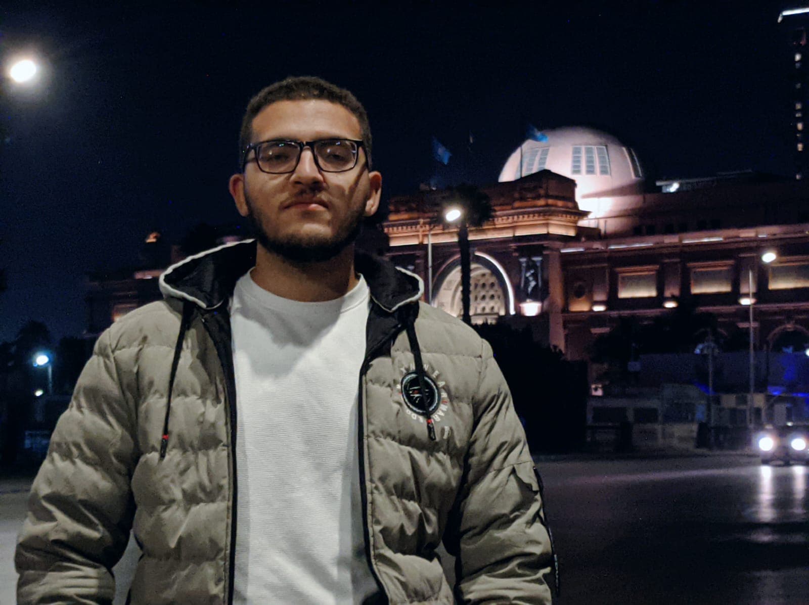 Bearded man in a puffer jacket posing before an illuminated domed building at night.