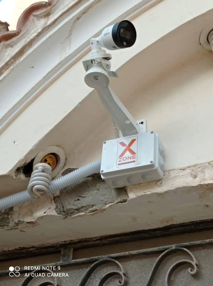 White security camera mounted on a wall above a junction box labeled X ZONE.