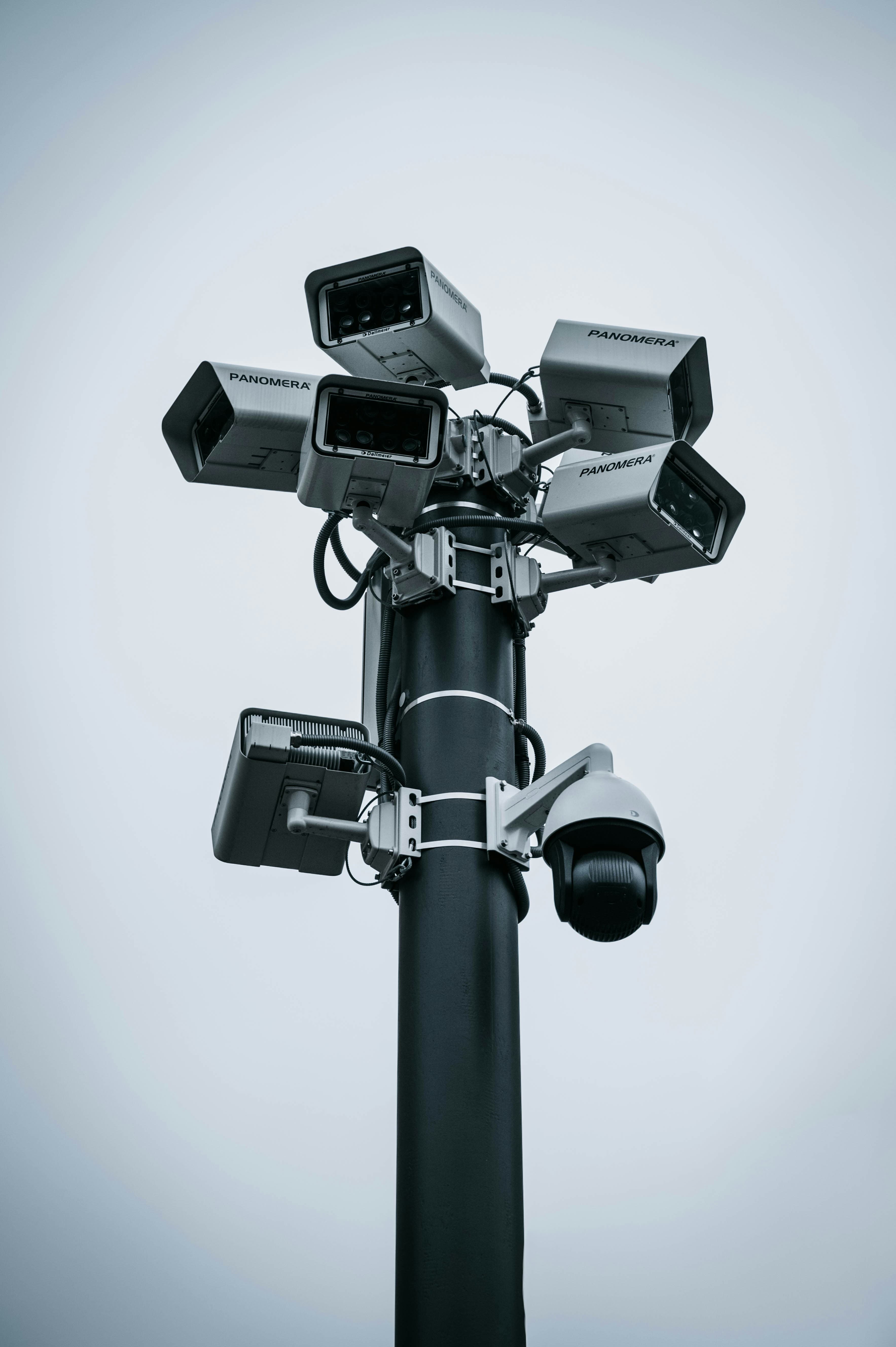 Multiple surveillance cameras mounted on a tall black pole against a plain gray sky.